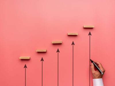 Businessman drawing upwards pointing arrows to support wooden pegs positioned in stairway like structure in a conceptual image of business growth and development.