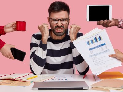 Outraged angry man has thick stubble, clenches fists in anger, unrecognizable people stretch hands to him with papers, mobile phone, notepad and mug, feels irritated with everybody, asks not bother