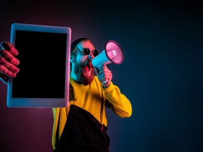 Showing tablet's blank screen. Caucasian man's portrait on gradient studio background in neon light. Beautiful male model with hipster style. Concept of human emotions, facial expression, sales, ad.