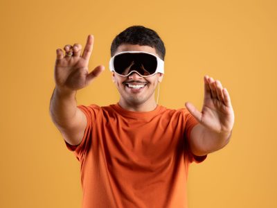 Modern imaging. Happy handsome brazilian man using modern vision pro glasses of virtual reality on yellow background, guy gesturing and smiling. Future gadgets technology concept